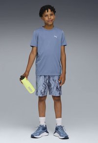 Μπλε αθλητική μπλούζα, μπλε σορτς με σχέδιο πλέγματος και μπλε αθλητικά παπούτσια. Κρατάει ένα φωτεινό κίτρινο παγούρι με μαύρο καπάκι.