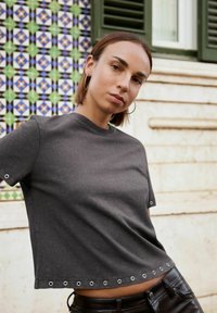 Žena s krátkými vlasy, oblečená v tmavě šedém tričku s průstřihy a černých kožených kalhotách, stojící proti obložené zdi a oknu.