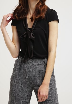 Femme portant un haut noir à manches courtes avec un nœud noué et un pantalon gris à carreaux taille haute sur un fond uni.