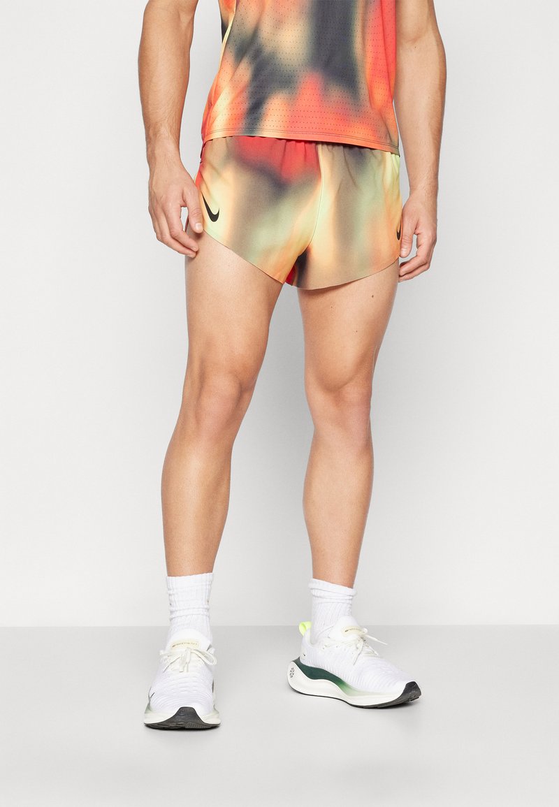 Shorts pour hommes en tie-dye orange, vert et jaune ; tissu léger, design ajusté. Associés à des baskets blanches et des chaussettes montantes.