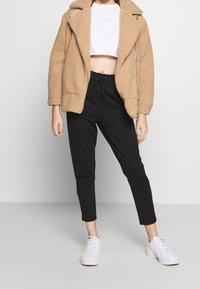 Beige fuskpälsjacka med dragkedja, avskuren vit t-shirt, svarta smala byxor och vita sneakers. Har mjuk textur och avslappnad design.