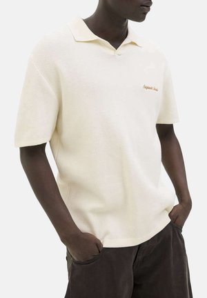 Homme portant un polo à manches courtes crème avec col et texte en écriture cursive sur la poitrine, les mains dans les poches d'un pantalon marron foncé.