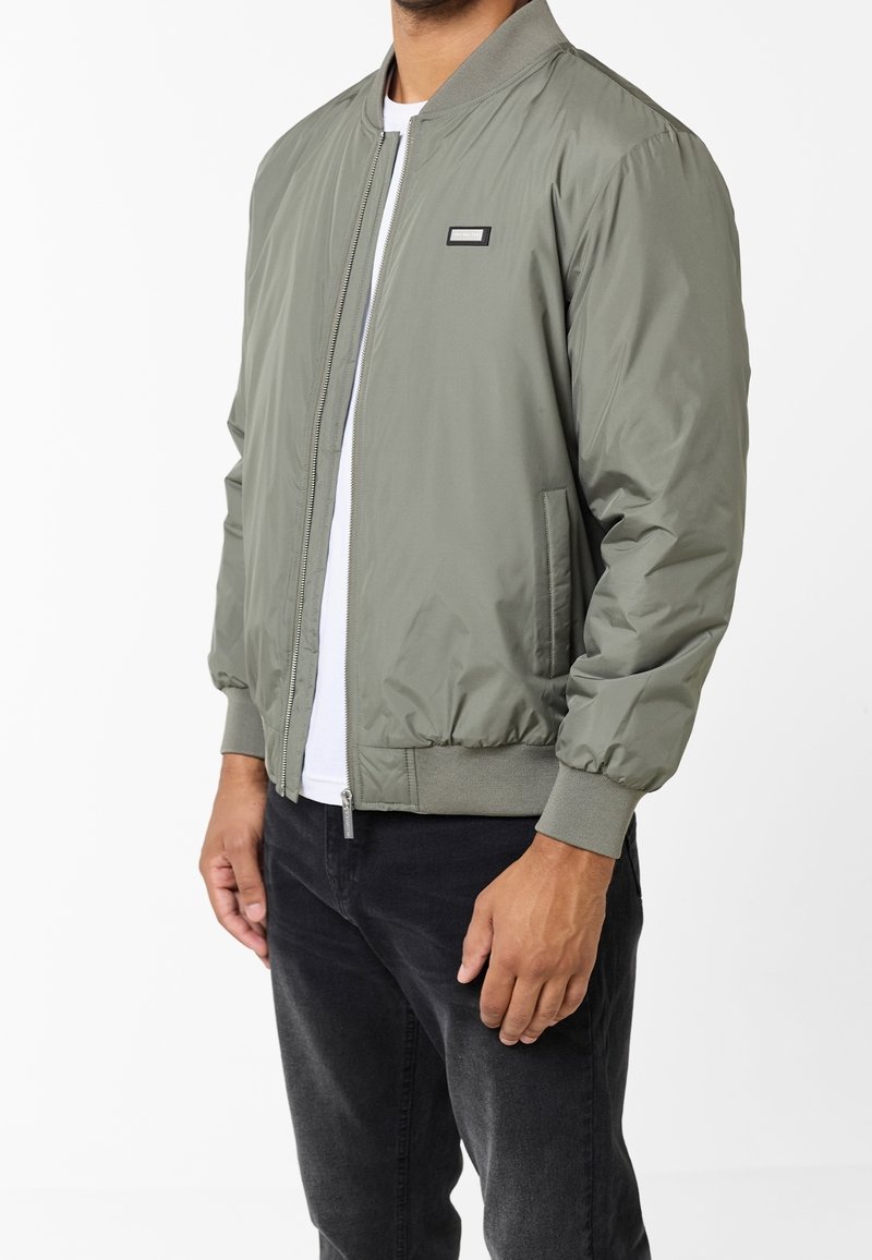 Homme portant une veste bomber vert olive clair avec col, poignets et ourlet côtelés, fermeture éclair devant et petit patch logo rectangulaire sur la poitrine.