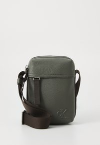 Hnedá textúrovaná kožená crossbody taška so strapcom z látky, s užším zipsom a embosovaným logom na prednej strane.
