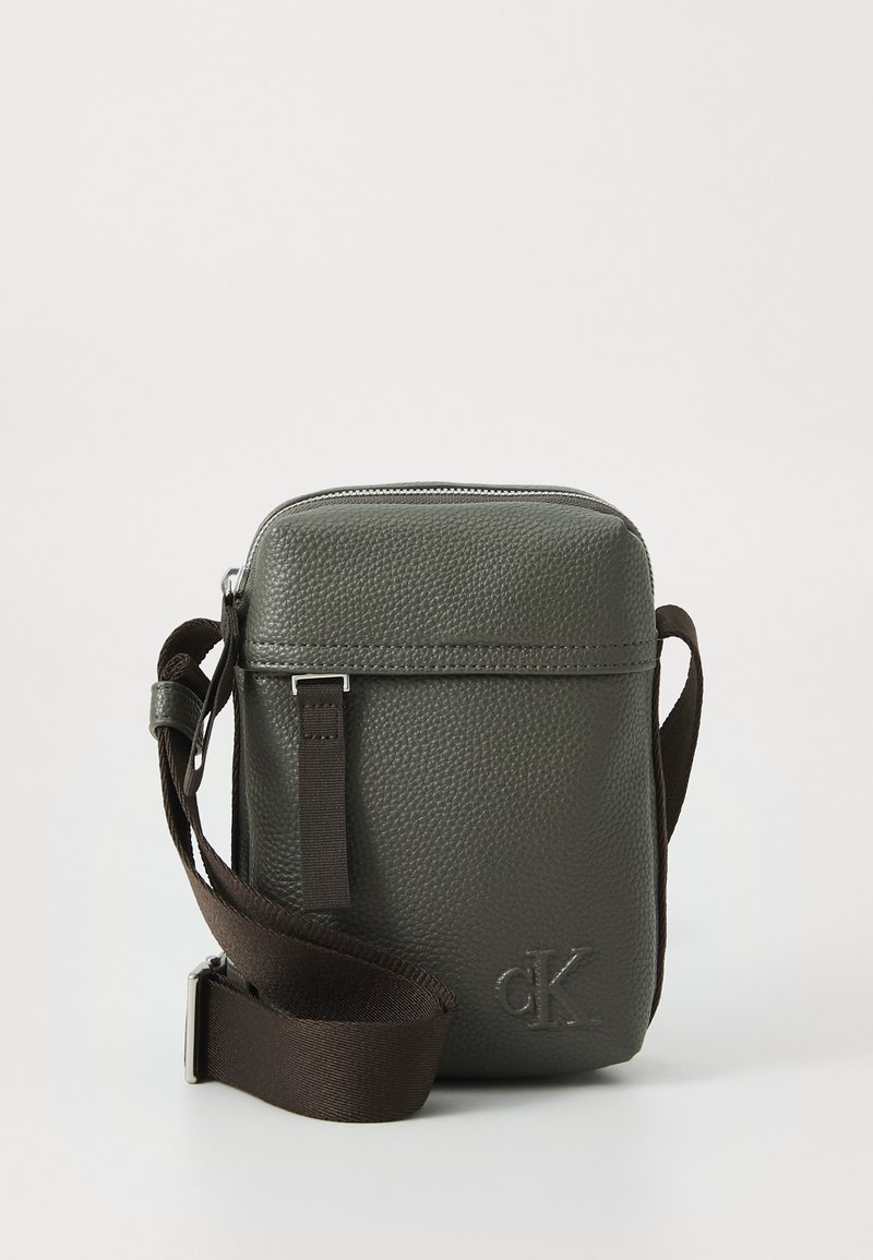 Hnedá textúrovaná kožená crossbody taška so strapcom z látky, s užším zipsom a embosovaným logom na prednej strane.