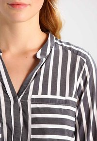 Blusa grigia e bianca a righe con collo a bottoni, dotata di una tasca sul lato sinistro del petto e una finitura di tessuto texturizzato.