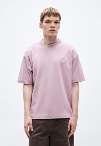 Svetloružové tričko s krátkym rukávom, okrúhlym výstrihom a textúrovanou okrúhlou vyšívanou záplatou na ľavej strane hrudníka. Jemný bavlnený materiál.
