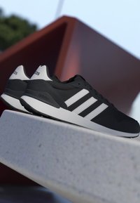 Sneakers Adidas in bianco e nero con tre strisce bianche, posizionate su una superficie di pietra contro uno sfondo esterno astratto.