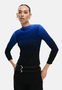 Top de manga larga azul y negra con un patrón de zigzag texturizado y un escote redondo, combinado con un cinturón negro de doble hebilla.