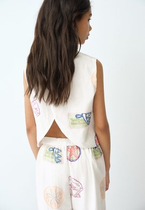 Fată cu păr lung și închis la culoare poartă un top alb fără mâneci și pantaloni cu petice brodate colorate, stând cu fața spre un perete alb.