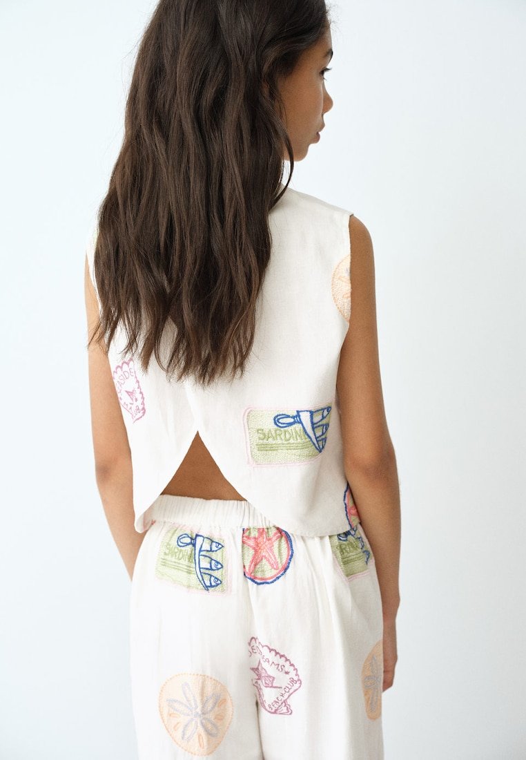 Fille aux longs cheveux foncés porte un haut blanc sans manches et un pantalon avec des patchs brodés colorés, debout face à un mur blanc.
