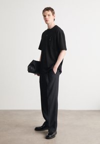 Černé tričko s krátkým rukávem a kapsou, kombinované s černými volnými kalhotami a lesklými černými botami, držící černou kabelku.