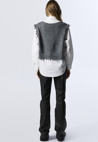 Maglione grigio corto con bordi sfilacciati, indossato sopra una camicia bianca. Jeans neri a campana con dettagli di cucitura bianca completano l'outfit.