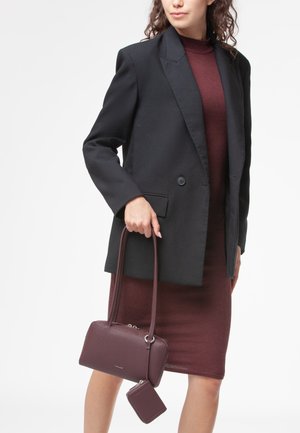 Blazer nero sopra un dolcevita bordeaux e una gonna a coste, mentre tiene una borsa a tracolla testurizzata color borgogna con un portafoglio abbinato.