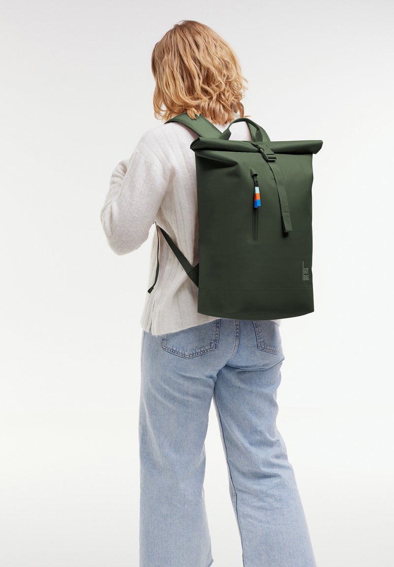 Mochila verde con tapa enrollable hecha de material duradero, que cuenta con un bolsillo frontal con cremallera, correas ajustables y un tirador de cremallera en bloques de color.