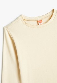 Haut à manches longues couleur crème avec une texture côtelée. Présente un col festonné et une petite étiquette sur le col. Tissu doux.