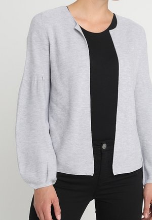 Hellgraue offene Strickjacke mit langen Puffärmeln, getragen über einem schwarzen Top und schwarzen Hosen bei einer Person, die vor einem neutralen Hintergrund steht.