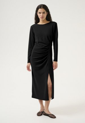 Black long-sleeve dress with a boat neckline, side ruching, and a thigh-high slit. Worn with black mesh slip-on shoes. Smooth texture.