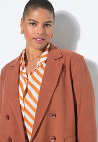 Hnědý lněný blejzr s dvojitým řádem knoflíků přes bílou košili s oranžovými vodorovnými pruhy. Jemná textura, střih na míru.