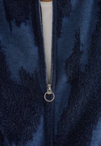 Marineblauer Zip-Pullover aus strukturiertem Material, mit einem samtigen, dunklen Muster auf der Vorderseite und einem silbernen Reißverschlussanhänger am Kragen.
