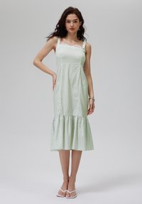 Robe verte claire avec des pois blancs. Présente un corsage ajusté, des fines bretelles, un bord en dentelle et un ourlet volanté évasé. Tissu lisse et doux.