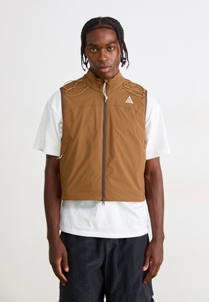 Brown, sleeveless vest with a high collar, front zipper, and adjustable cord detailing. Worn over a white shirt. Features logo on chest.