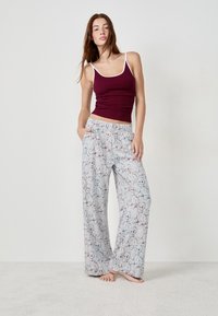 Femme debout pieds nus, portant un débardeur bordeaux et un pantalon de pyjama gris ample avec des illustrations de teddy bears sur un fond uni.