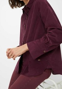 Chemise en velours côtelé bordeaux avec un devant boutonné, des manches longues avec des détails froncés et un ourlet incurvé. Accents en métal doré visibles.