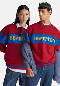 Czerwone i niebieskie bluzy z napisem „BENETTON”, z kołnierzykiem i paskami, noszone na codziennym dżinsie. Gładkie tło podkreśla wzór.