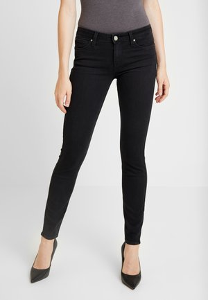 Žena v přiléhavých černých džínách, šedém tílku a černých lodičkách s špičatou špičkou, stojící před jednoduchým světlým pozadím.