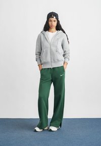 Sivá mikina na zips s prednými vreckami, skombinovaná s voľnými zelenými teplákmi s bielym logom Nike. Biele tenisky dotvárajú vzhľad.