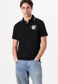 Schwarzes Poloshirt mit weißen Akzenten, kurzen Ärmeln und einem Logo auf der Vorderseite. Kombiniert mit hellblauen Jeans, die Taschen und einen lässigen Schnitt haben.