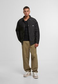 Fekete, texturált dzseki zsebbel és gombos zárással, fekete trikó fölött viselve. A szettet világos oliva nadrág és fehér sportcipő egészíti ki.