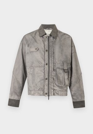 Man wearing gray jacket, white shirt, black wide-leg jeans, black sneakers, and black sunglasses, standing with one hand in pocket.