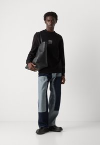 Svart genser med logo-lapp, kombinert med lappede jeans i lys og mørk denim, og en stor svart lær totebag. Svart fottøy.