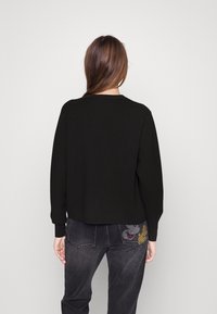 Czarny sweter z długim rękawem o luźnym kroju. Wykonany z bawełny. Zestawiony z szarymi dżinsami z kolorową haftowaną aplikacją na tylnej kieszeni.