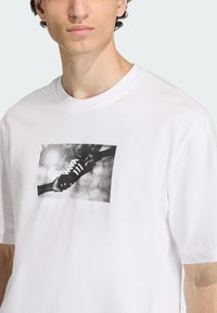 Λευκό βαμβακερό μπλουζάκι με γράφημα που απεικονίζει ένα χέρι να κρατάει ένα μαύρο αθλητικό παπούτσι μπροστά σε ένα συννεφιασμένο φόντο, με στρογγυλή λαιμόκοψη και κοντά μανίκια.