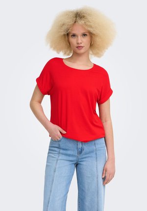 Giovane donna con capelli biondi ricci che indossa una maglietta rossa a maniche corte e jeans azzurri a vita alta, in piedi con una mano in tasca.