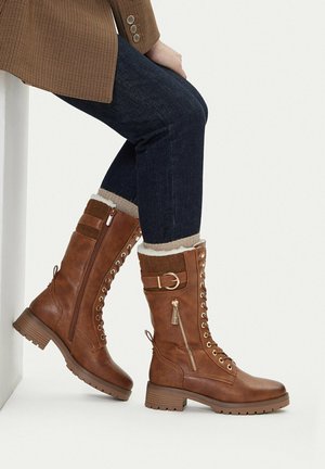 Person trägt braune Leder-Schnürstiefel mit seitlichen Reißverschlüssen und Schnallen, dunkle Jeans und eine braun karierte Jacke, sitzt mit übereinandergeschlagenen Beinen.