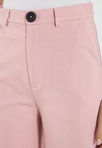 Pink tailored trousers made of smooth fabric; features a front button, belt loops, and side pockets with subtle stitching details.
