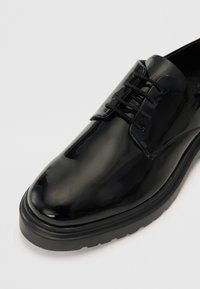 Chaussure en cuir verni noir avec une surface lisse et brillante, bout rond, lacets noirs, et une semelle en caoutchouc épaisse et texturée.