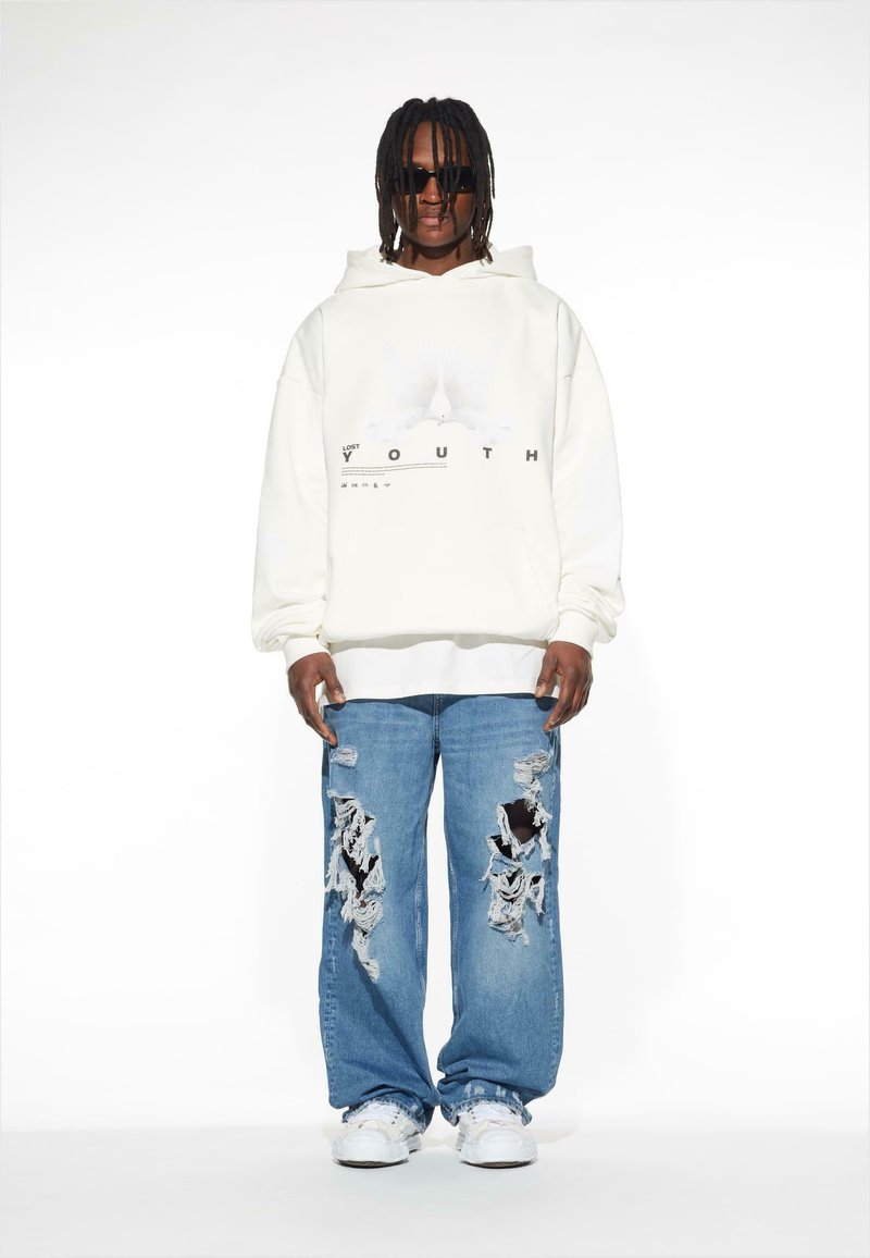 Felpa bianca con stampa grafica, jeans blu denim larghi con grandi strappi e sneakers bianche. Caratterizza una vestibilità comoda e dettagli di cucitura visibili.