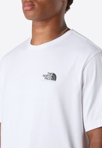 Nærbillede af en person iført en hvid kortærmet T-shirt med The North Face's sorte logo og slogan på brystet, samt en sølv kæderekæde.