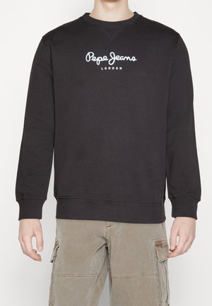 Osoba ubrana w czarną bluzę Pepe Jeans London i beżowe spodnie typu cargo, stojąca na tle jednolitego białego tła.