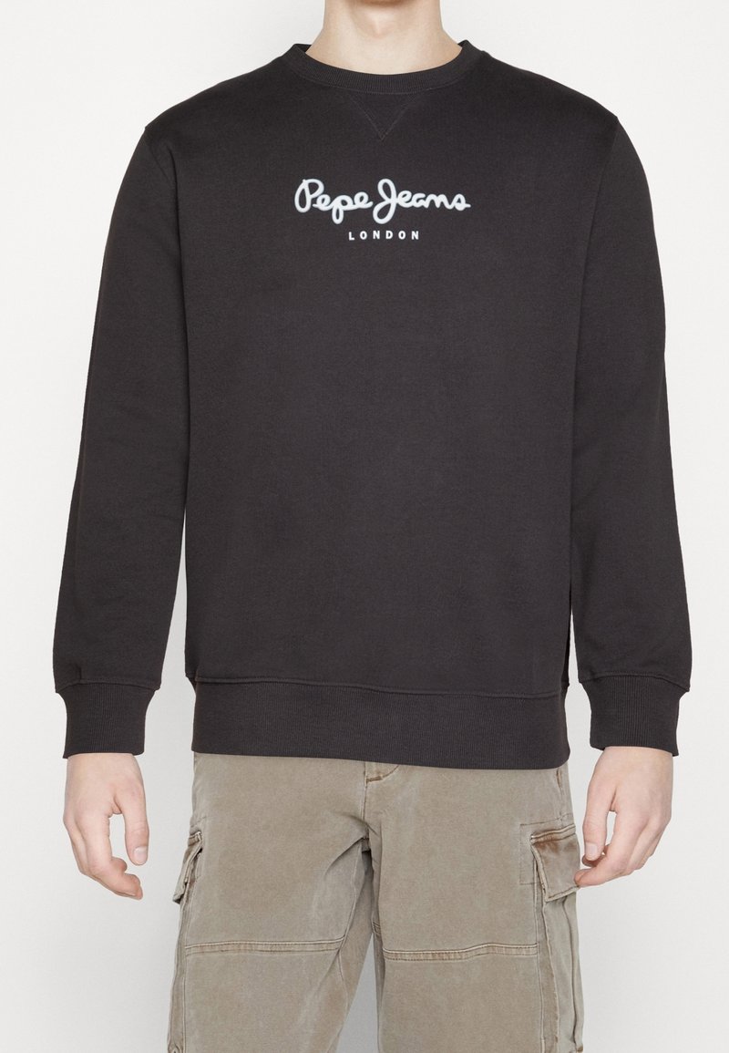 Personne portant un sweat-shirt noir Pepe Jeans London et un pantalon cargo beige, debout devant un fond blanc uni.