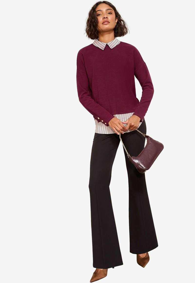Maglione bordeaux sopra una camicia a righe bianca e nere, abbinato a pantaloni neri svasati. Una piccola borsa bordeaux con tracolla a catena.