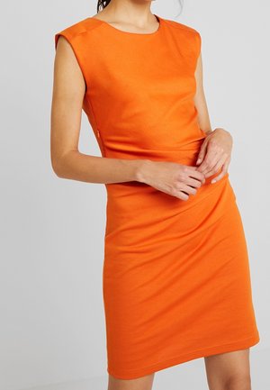 Frau trägt ein enganliegendes, ärmelloses, leuchtend oranges Kleid und steht vor einem schlichten hellgrauen Hintergrund.
