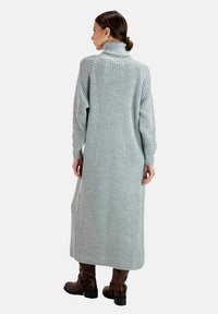 Langes, geripptes Strickkleid aus hellgrauem Wollmix mit hohem Kragen und langen Ärmeln, ergänzt durch knielange braune Lederstiefel.