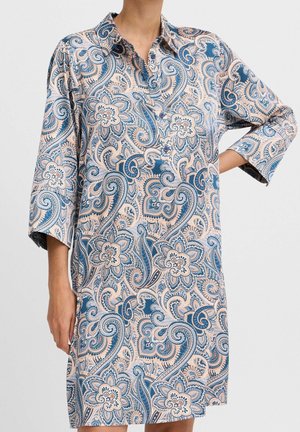 Kvinde iført en blå og beige paisley blomsterprintet skjortekjole med lange ærmer, kort krave og knapper.