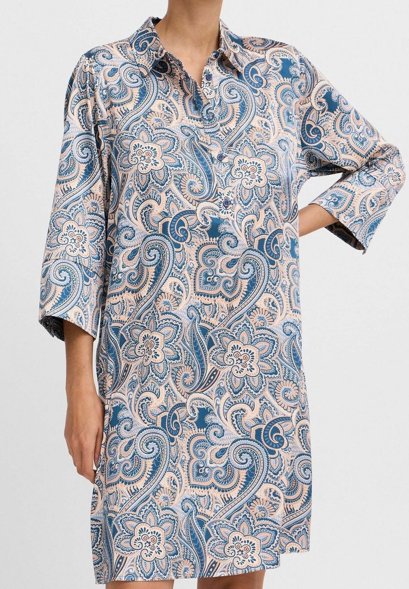 Femme portant une robe-chemise à manches longues à motifs floraux paisley bleu et beige, avec un petit col et des boutons.